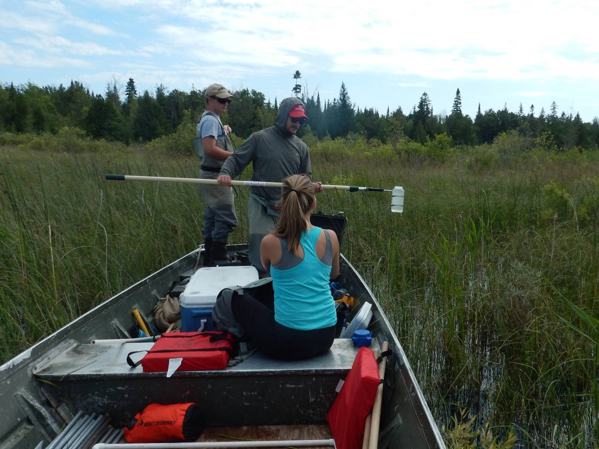 Three technicians on the jonboat collect a water sample using a bottle on a long stick.